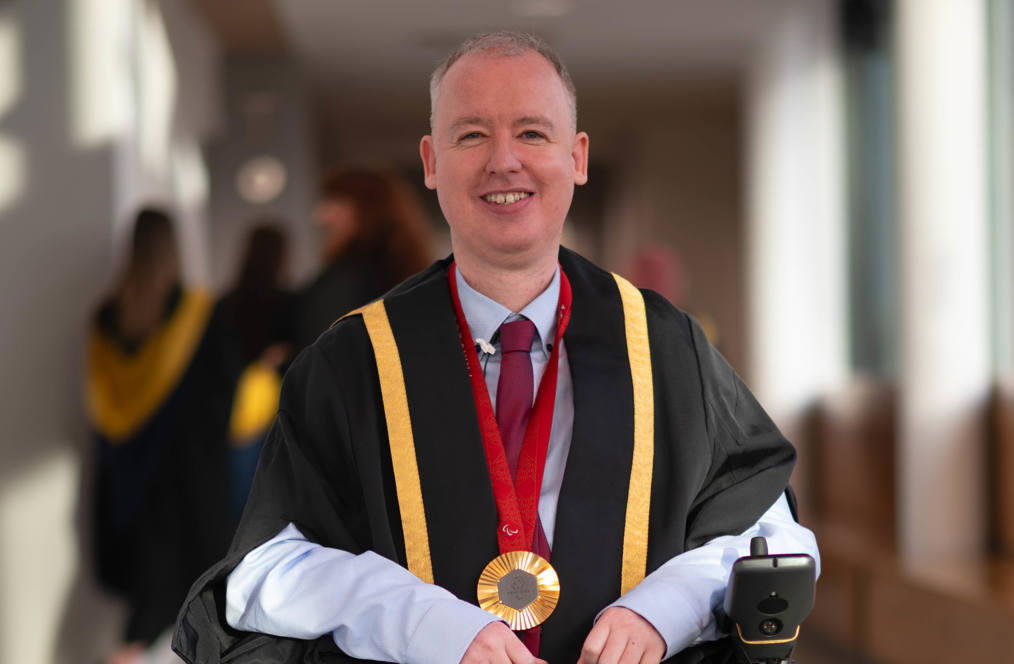 Stephen McGuire, a Paralympic gold medallist, wearing formal graduation attire with gold trim and a medal around his neck, seated in a wheelchair. He is smiling and positioned in a well-lit hallway, with blurred figures of graduates in the background.